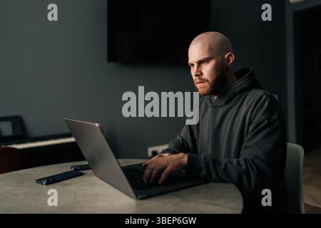 Focused male developer in casual hoodie typing code on laptop computer, developing software in dark home office, looking to screen with serious Stock Photo