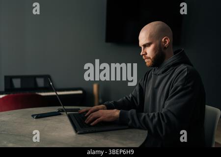 Side view of focused male programmer wearing dark hoodie typing code on laptop computer, sitting at table near window in contemporary urban apartment. Stock Photo