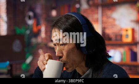 CTO manager in artificial intelligence focused startup drinking coffee, listening music and debugging applications. Asian technician putting headphones on, doing code testing, camera B close up Stock Photo