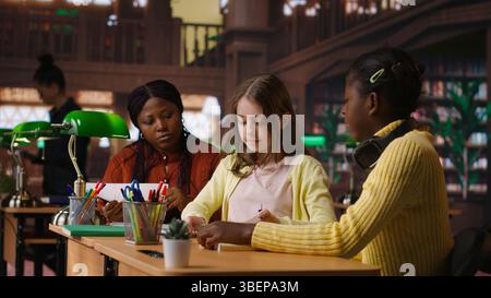 Preteens girls and their professor collaborating on schoolwork at the library, focused on academic materials to write a good essay. Students collect extra information with private lessons. Camera B. Stock Photo