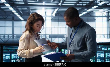 Team of data center supervisors reading paperwork files, reviewing system diagnostics. Server room coworkers looking over maintenance schedules documents and cybersecurity protocols Stock Photo