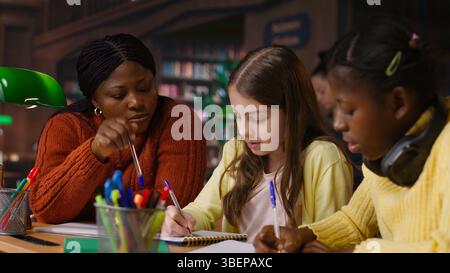 Female teacher assisting pupils with class homework and mentoring, providing academic instruction and support to help pass school assignments. Tutor lecturer gives private lessons. Camera B. Stock Photo