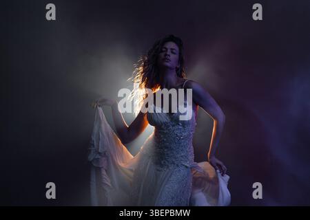 Graceful dancer in flowing white gown, silhouetted by mysterious backlight in moody atmosphere Stock Photo