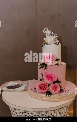 A beautifully decorated two-tier blush pink wedding cake adorned with white and pink roses and a gold "Mrs" cake topper sits on a circular display tab Stock Photo