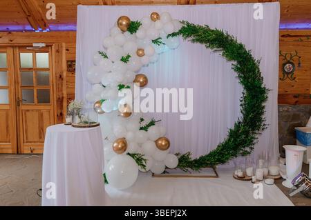 Elegant wedding backdrop featuring a white fabric drape adorned with a lush balloon garland in shades of white and gold, complemented by vibrant green Stock Photo
