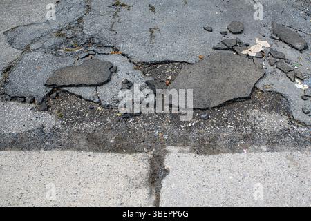 Broken asphalt at the side of a road, next to a concrete sidewalk. Closeup view, only part of the road and sidewalk visible. Stock Photo