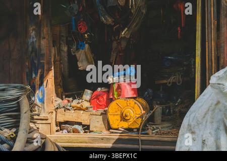 The dimly lit interior of a workshop or storage area is filled with an assortment of industrial items, including cables, ropes, metal parts, a large y Stock Photo