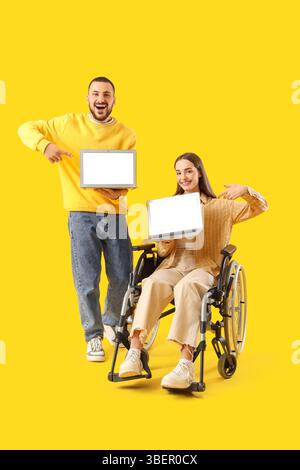Young woman in wheelchair, with husband and laptops on yellow background Stock Photo