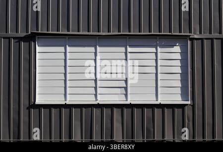 Rectangular window with white louvers louvres on a gray grey corrugated metal wall Stock Photo