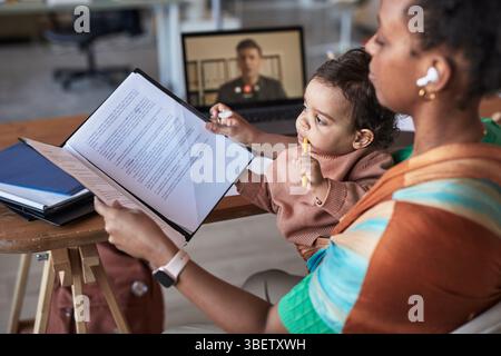 Woman multitasking with busy workload, holding child while participating in video call. Child sitting on mother's lap observing laptop screen, contrib Stock Photo
