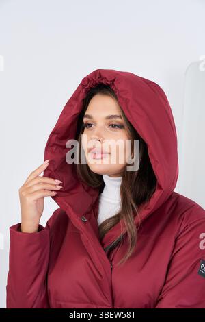 Woman with long hair wearing a red hooded jacket and a white turtleneck. She holds the hood with one hand. Her expression is calm and thoughtful Stock Photo
