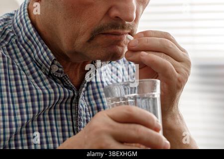 Close-up of a senior man in a plaid shirt taking a medication pill with a glass of water. Stock Photo