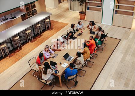 Top-view of a group of diverse creative professionals seated around a long table in a modern office environment, using laptops and discussing ideas ov Stock Photo