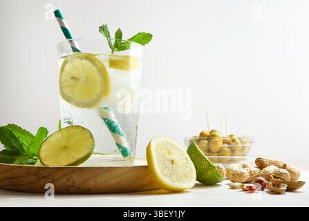 Refreshing carbonated lemon drink on a wooden table with snacks such as peanuts and olives decorated with mint leaves and lime and lemon slices with w Stock Photo