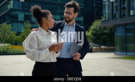 African American woman businesswoman female girl in city walking using mobile phone chat Caucasian man businessman guy male approach walking to lady Stock Photo