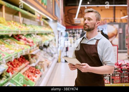 Store employee checking inventory using tablet, standing by fresh produce aisle. Focused worker engaging in tasks in well-lit grocery store environmen Stock Photo