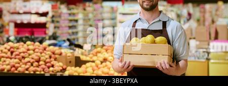 Website banner of worker holding wooden box filled with yellow peppers while standing in grocery store smiling behind fruit displays Stock Photo