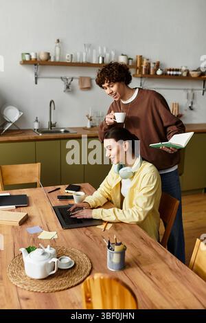 A man playfully looks over a womans shoulder as she focuses on her laptop at their dining table. Stock Photo