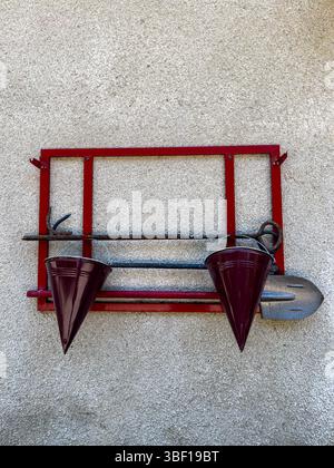 Firefighting equipment with red metal frame, buckets, shovel and hook hanging on textured wall, close-up, daylight, safety and emergency concept. Stock Photo