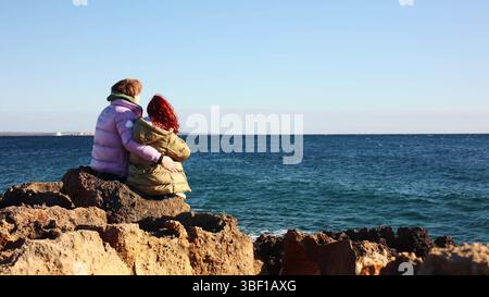 a loving couple sits on the seashore hugging. happy family spending time with each other enjoying vacation at seaside It's spring, people are wearing Stock Photo