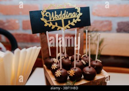 A close-up photo of a table setting with a wooden stand holding a collection of cake pops. The cake pops are dipped in chocolate and decorated with sm Stock Photo