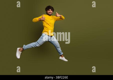 Young man in vibrant yellow shirt jumps joyfully with a thumbs-up on khaki backdrop Stock Photo