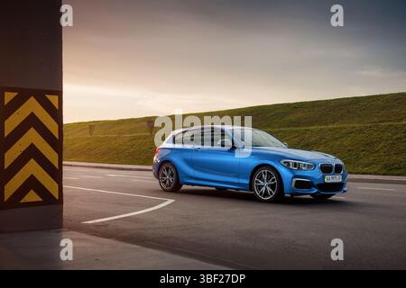 blue BMW M140i in a parking lot at sunset. Three quarter front side view of sporty BMW 1-series (F21) hatchback with striped pillar on the left. Stock Photo