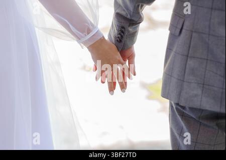 A close-up shot featuring a bride and groom holding hands, emphasizing their wedding rings and elegant outfits, set against a soft-focus, snowy outdoo Stock Photo