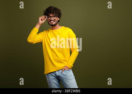 Young man in yellow shirt smiling confidently and adjusting glasses against a green backdrop for casual lifestyle themes Stock Photo