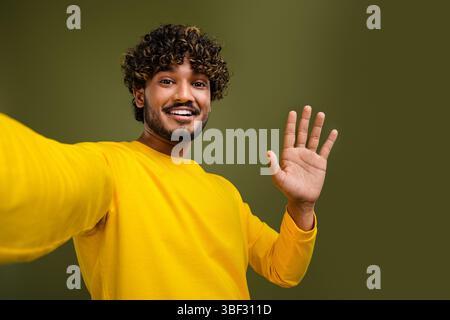 Young man with curly hair in yellow shirt waving hand on khaki background, friendly gesture, modern casual style Stock Photo