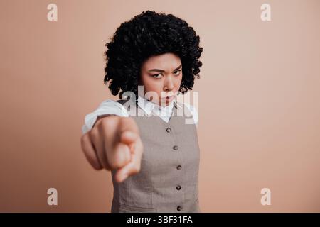 Confident businesswoman with curly hair pointing forward in formal attire against beige background Stock Photo