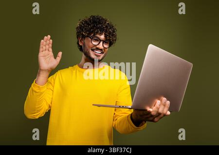 Young man in a yellow shirt waving while using a laptop, smiling, against a khaki background, promoting online engagement Stock Photo