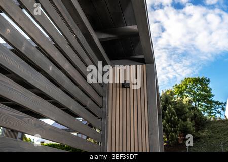 Sunlight casts shadows on the sleek wooden slats of a modern structure outside a home, surrounded by lush greenery and a bright sky. Stock Photo