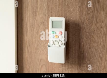 White remote control in a wall holder mounted on a wooden surface, used for air conditioning or home devices. Stock Photo