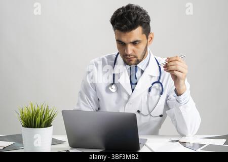 Serious arab practitioner working at computer. Handsome physician in white medical gown with stethoscope sitting at desk with laptop. Professional med Stock Photo