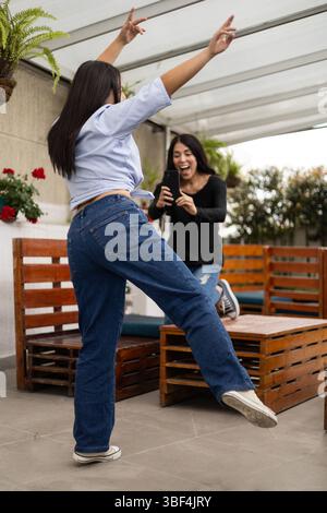 Two women share joyful moments taking pictures on a wooden patio with tables, blending friendship, outdoor relaxation, and casual style under natural Stock Photo