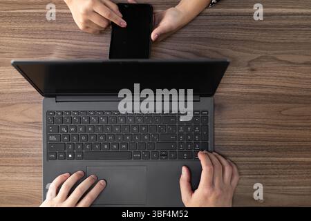 Dynamic overhead shot captures hands skillfully managing laptop and smartphone, reflecting modern multitasking and seamless digital connectivity. Stock Photo
