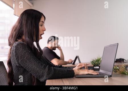 Woman looks surprised at her laptop screen while man works in the background. A candid moment capturing reaction, emotion, and real-time discovery. Stock Photo