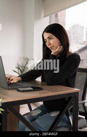 Young woman stares at laptop in deep thought while working alone in a modern office. A moment of focus, analysis and professional reflection. Stock Photo