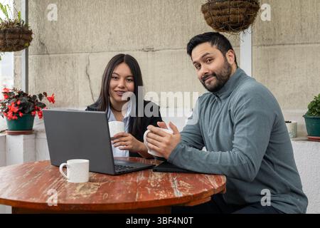 Two coworkers smiling while working on laptops and drinking coffee in a relaxed outdoor workspace. Casual, friendly and collaborative scene. Stock Photo