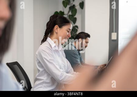 Collaborative work session with two women and one man sharing a long desk, using laptops and a tablet in a modern office space. Stock Photo