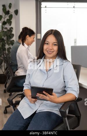 A cheerful woman works on her tablet while seated on a swivel chair. Her teammates are in the background of a bright modern office. Stock Photo