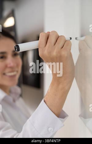 Smiling confidently, a young woman writes on a whiteboard while presenting herself to her new team during an onboarding session. Stock Photo