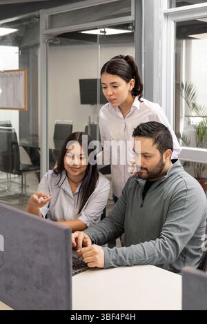 Three coworkers gathered around a computer, engaged in teamwork and problem-solving in a bright, modern office space. Stock Photo