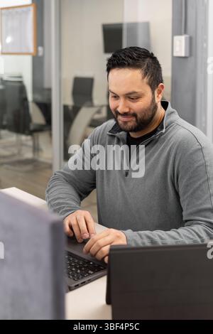 A man with a beard wearing a gray blazer types on his laptop at a desk. He is concentrated and working in a bright office space, suggesting productivi Stock Photo