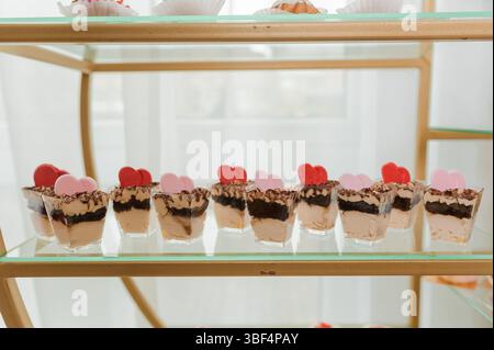 A close-up shot showcases a golden metal shelf displaying a row of chocolate dessert cups adorned with pink and red heart-shaped toppers, ideal for Va Stock Photo