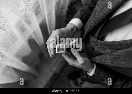 A close-up shot of a groom wearing a gray tweed suit carefully fastening a silver rectangular watch on his wrist, showcasing a sophisticated wedding p Stock Photo