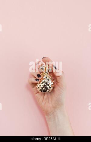 Creative top view flat lay of woman hand with black manicure holding small golden pineapple with copy space on pink paper background in minimalism sty Stock Photo