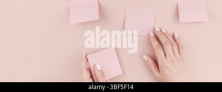 Long wide banner of workspace desk with millennial pink sticker paper sticky note mock up in woman hands holding and gluing them on wall with copy space b Stock Photo