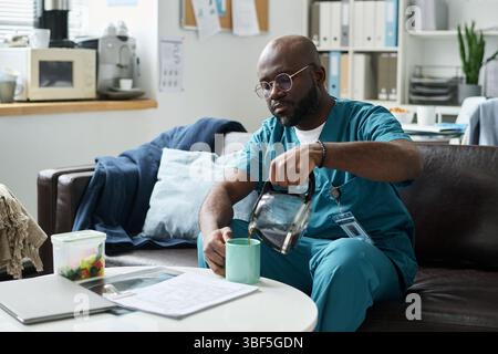 African American male doctor sitting in office with medical documents and drink. He is wearing scrubs and glasses while carefully pouring water into his cup Stock Photo